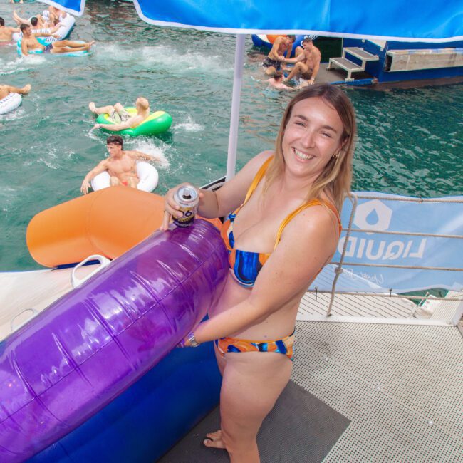 A smiling woman in a colorful bikini holds a large purple pool float on a dock by the water, with people swimming and relaxing on inflatables in the background.