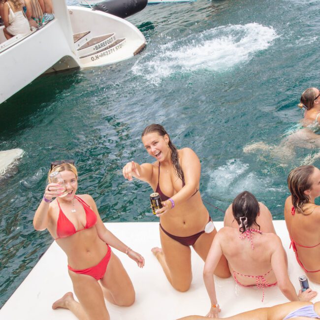 A group of women in swimsuits enjoy drinks on a floating platform in the ocean near boats, smiling at the camera while others swim and relax in the water.