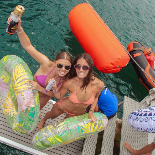 Two smiling women in sunglasses and bikinis stand on a dock holding drinks and colorful pool floats, with green water and inflatable rafts in the background on a sunny day.