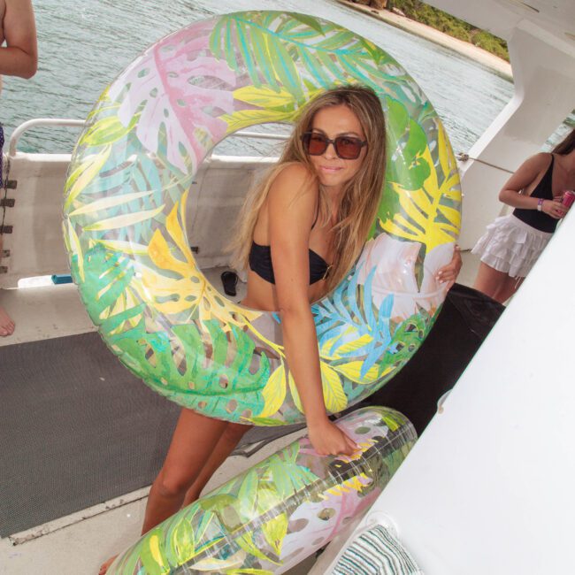A woman in sunglasses and a black swimsuit stands on a boat holding two inflatable pool rings with tropical leaf patterns. She poses playfully, with water and a shoreline visible in the background.