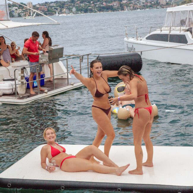 Three women in bikinis smile and pose on a floating platform in the water near boats with people socializing. The scene is festive, with city buildings and greenery visible in the background.