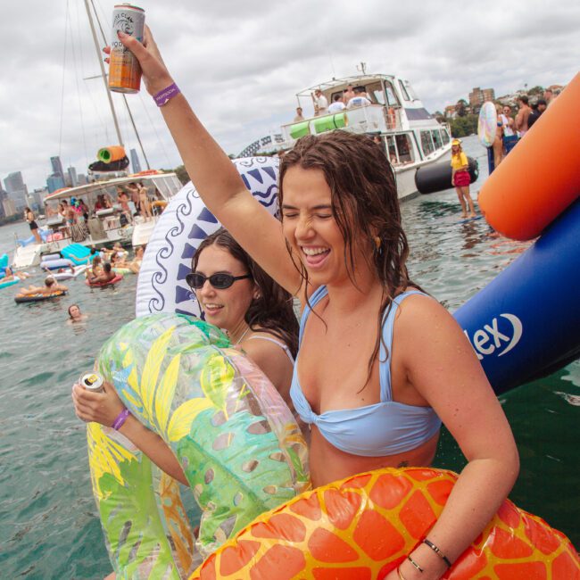 Two young women in swimsuits smile and raise drinks while floating on colorful inflatables at a lively party on the water, surrounded by other people, boats, and the city skyline in the background.