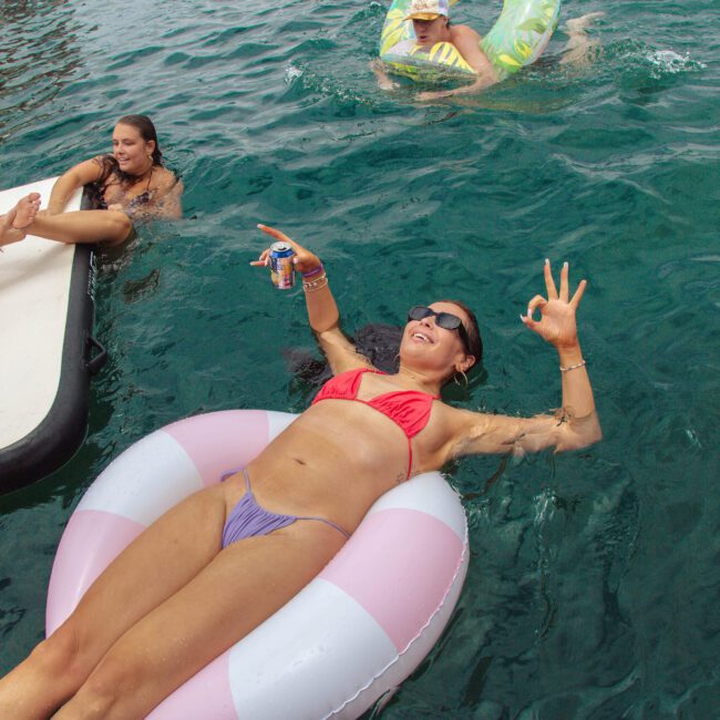 A woman in a bikini floats on a pink and white pool ring in clear water, making an "OK" hand gesture and holding a drink. Others swim and relax nearby. The atmosphere is relaxed and summery.