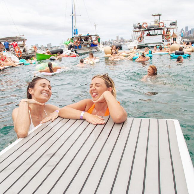 Two women smile and pose at the edge of a floating dock in the water, surrounded by people on inflatables and boats, with a city skyline visible in the background under a cloudy sky.