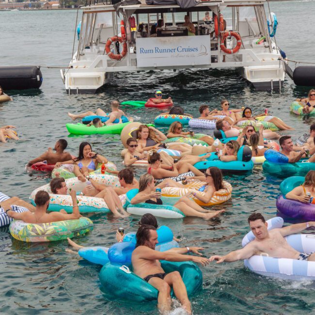Dozens of people float on colorful inflatables in the water near a large boat that reads “Rum Runner Cruises.” The scene is lively and festive, with people relaxing and interacting.