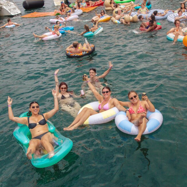 A group of people relax on colorful pool floats in the water, smiling and making peace signs, while others float and swim nearby during a lively outdoor gathering.
