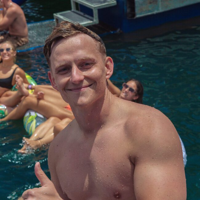 A smiling man with wet hair gives a thumbs up while standing in the water near a group of people relaxing on pool floats at a sunny outdoor event. A logo in the bottom right reads "Yacht Social Club.