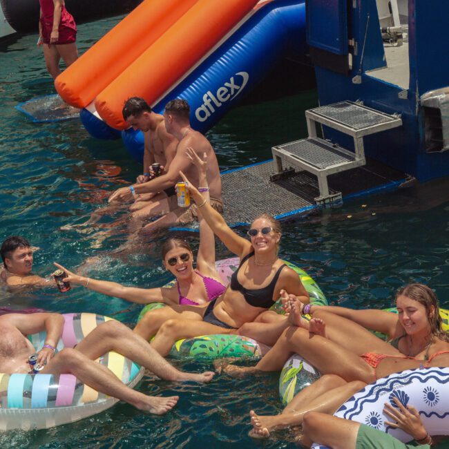 A group of people relax on colorful pool floats in the water near a dock, smiling and raising their arms in celebration. Others sit at the edge of the dock, dipping their feet in, with inflatable slides in the background.