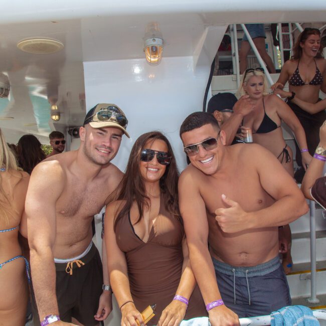 A group of young adults in swimsuits smile and pose on a crowded boat party. Some wear sunglasses and wristbands. Other people mingle and chat in the background near stairs. The atmosphere is sunny and lively.