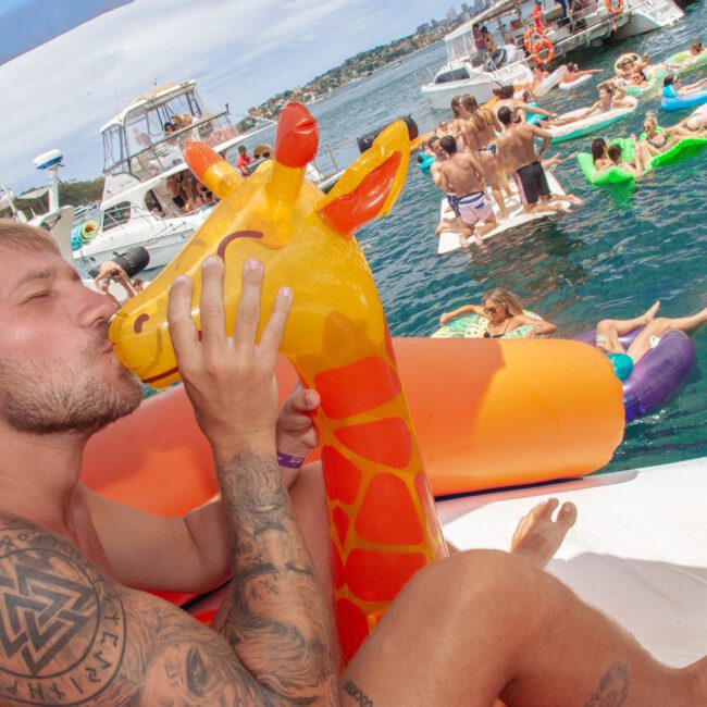 A man with tattoos kisses a large inflatable giraffe on a boat, while people on floats and boats enjoy a sunny day on the water in the background.