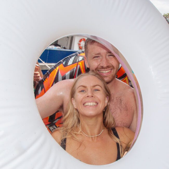 A smiling woman and a shirtless smiling man pose together, framed by the center of a large white pool float. The background shows a bright, sunny outdoor setting.