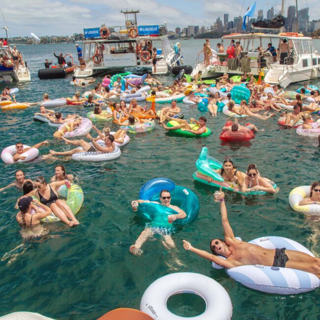 A large group of people float in colorful inflatable tubes and loungers in a bay, surrounded by anchored boats, with a city skyline in the background under a sunny sky.
