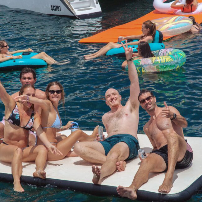 A group of five people smile and pose on a floating mat in a lake, surrounded by others relaxing on pool floats and inflatable rafts, with boats in the background on a sunny day.