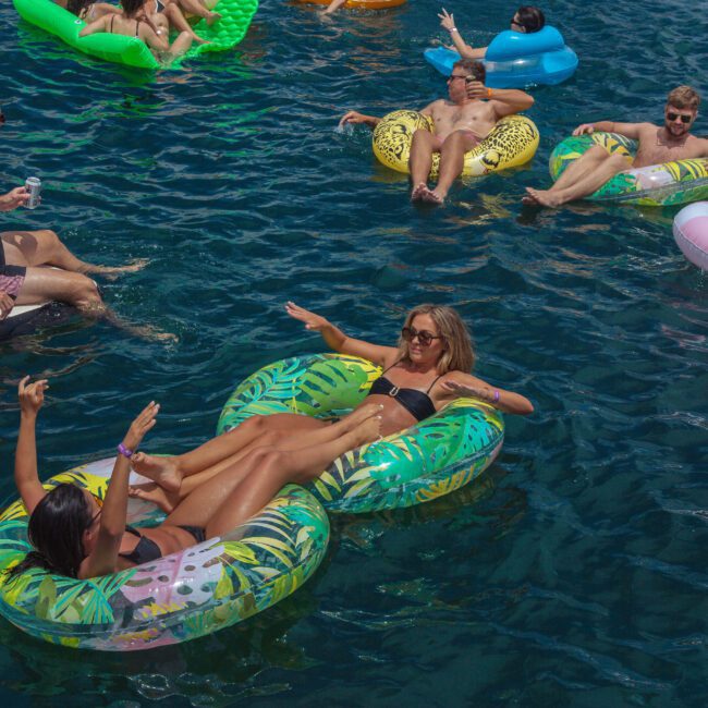 People relax on colorful inflatable floats in deep blue water during a sunny day. Some smile and wave, while others lounge casually. The mood is lively and social, with a “Yacht Social Club” logo in the bottom right corner.