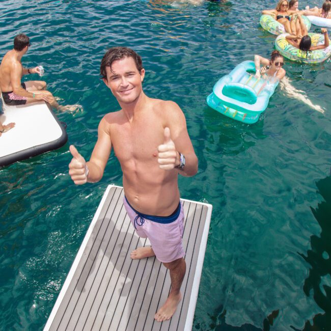 A man in pink swim trunks stands on a floating platform in the water, smiling and giving two thumbs up. Around him, people relax on inflatable floats and platforms, enjoying a sunny day at a pool party.