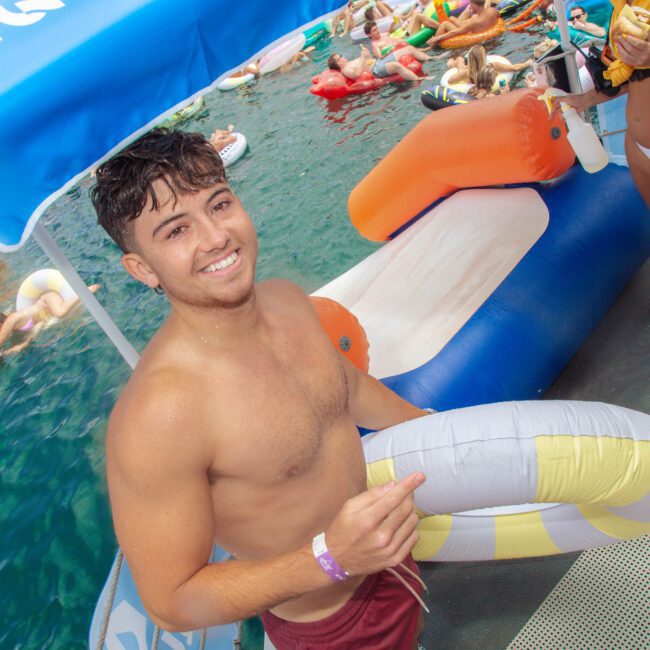 A young man in swim trunks smiles while holding an inflatable ring on a dock by the water, surrounded by people on floats at a lively pool or lake party.