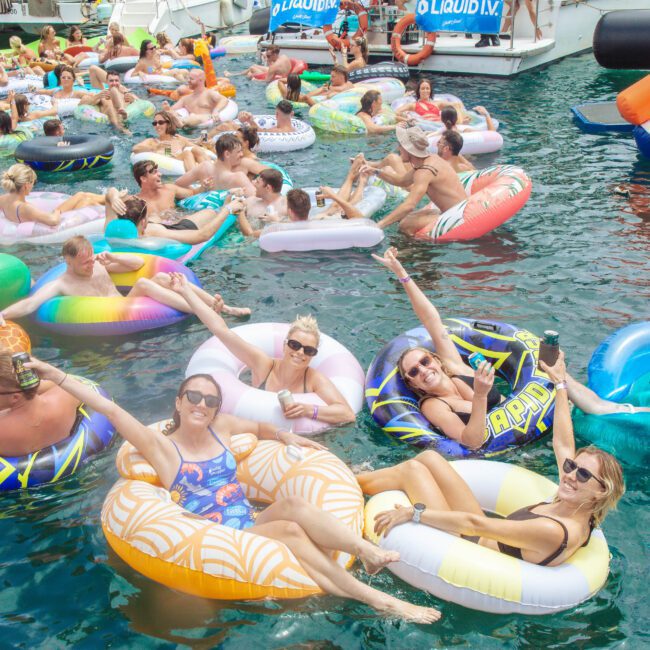 A large group of people relax on colorful inflatable floaties in the water, holding drinks and smiling at a lively pool party with boats in the background.
