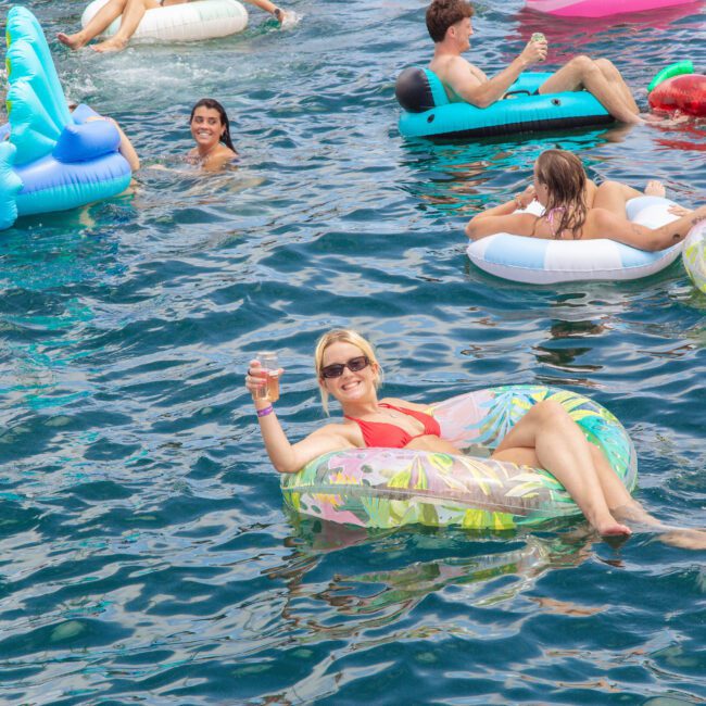 A woman in sunglasses smiles and holds a drink while lounging on an inflatable float in a pool, surrounded by others on colorful floats, enjoying a sunny day on the water.