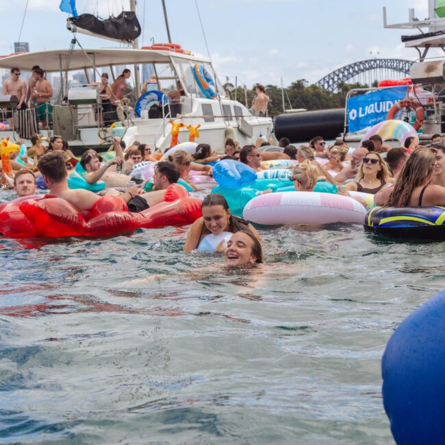 A large group of people float on inflatable tubes and swim in the water at a lively outdoor pool party, with boats, banners, and a city skyline visible in the background.