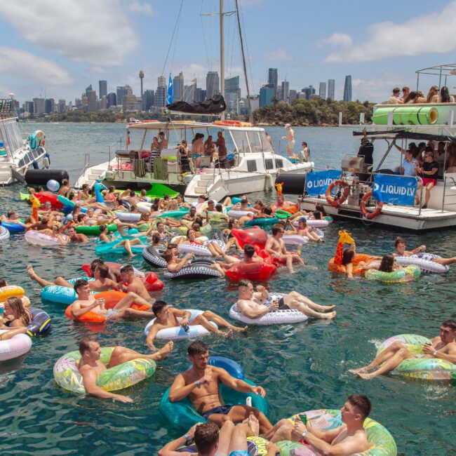A crowd of people relax on colorful inflatable pool floats in the water near anchored boats, with a city skyline in the background under a partly cloudy sky.