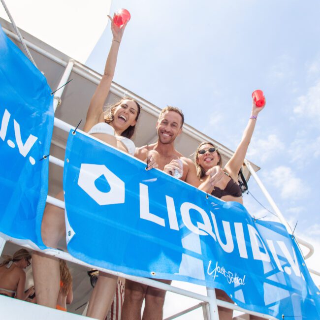 Three people in swimsuits smile and raise drinks while standing on a boat deck behind blue "LIQUID I.V." banners, enjoying a sunny day with a bright blue sky in the background.