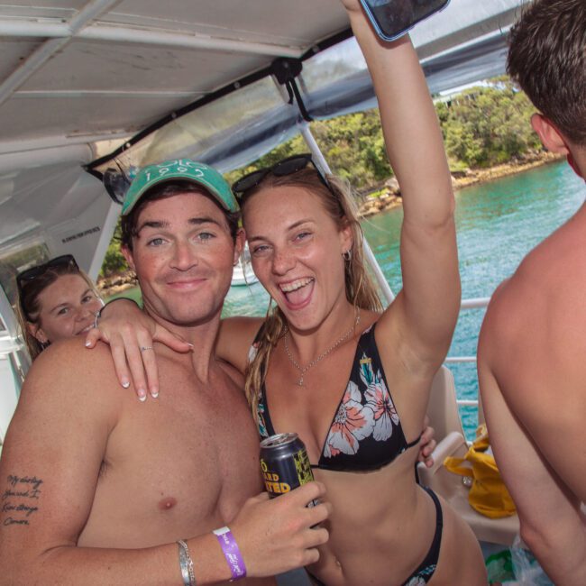 Two young adults in swimsuits smile and pose together on a boat, with one holding a can and raising an arm. Other people are nearby; water and trees are visible outside. The atmosphere is relaxed and festive.