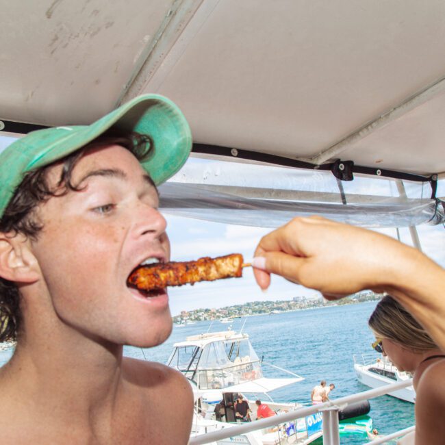 A man in a green cap eagerly eats grilled food from a skewer held by another person, while sitting on a boat with water and other boats visible in the background. Two other people are nearby, one looking at the camera.