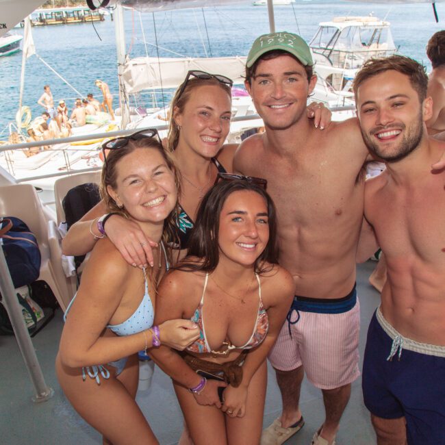 Five young adults in swimwear smile and pose together on a boat, with other people, boats, and water visible in the sunny background. One person is holding a can, and everyone appears to be enjoying a lively day.