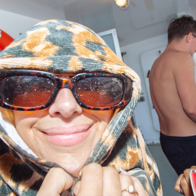 A person smiles at the camera, wearing sunglasses and covering their head with a patterned towel. Other people stand on a boat deck in the background near a life preserver.