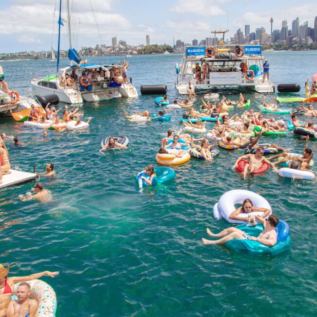 A lively group of people enjoy a floating party in a harbor, relaxing on colorful inflatables in the water near docked boats, with a city skyline in the background on a sunny day.