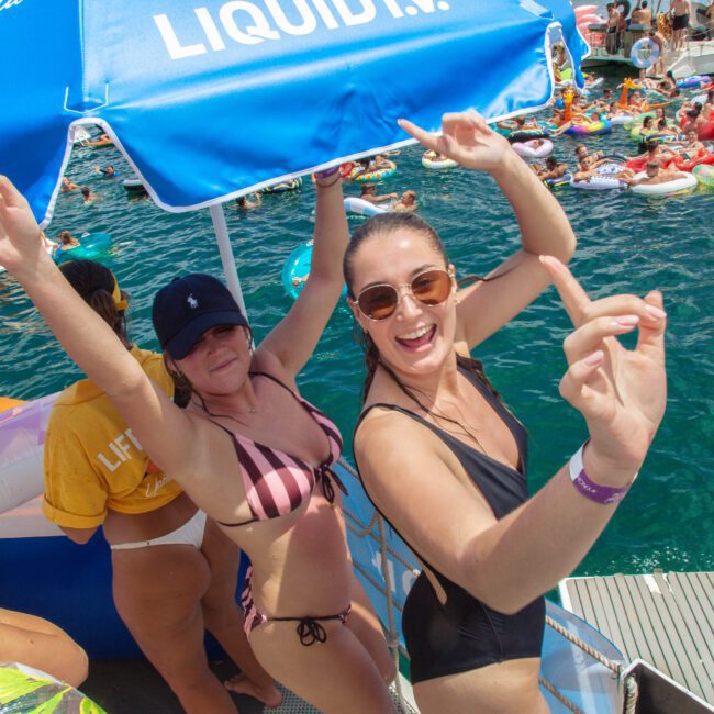 Two women in swimsuits pose and smile under a blue umbrella on a boat during a lively pool party, with people on colorful floaties in the water behind them. The atmosphere is festive and joyful.