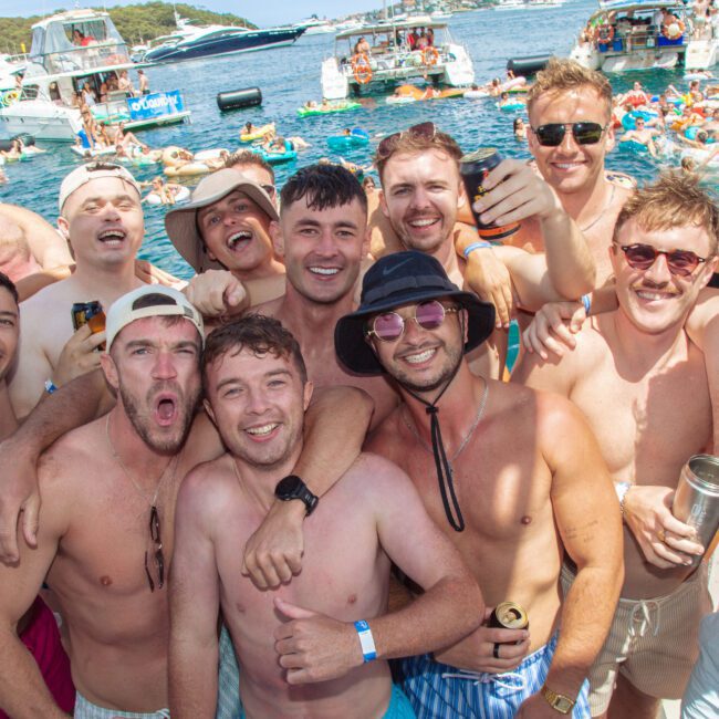 A group of smiling young men in swimwear pose together on a boat with drinks in hand, surrounded by other people, boats, and inflatables on a sunny day at a lively water party.