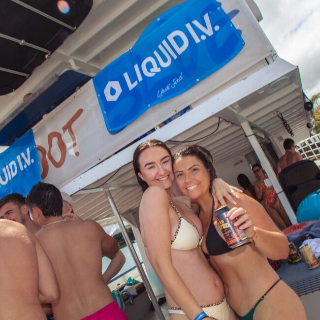 Two women in bikinis smiling and posing for a photo on a boat surrounded by other people. Behind them are banners that read "Liquid I.V." The atmosphere is festive and sunny near the water.