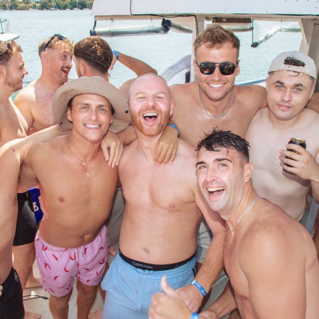A group of smiling, shirtless young men pose together on a boat during a sunny day, holding drinks with water and another boat visible in the background.