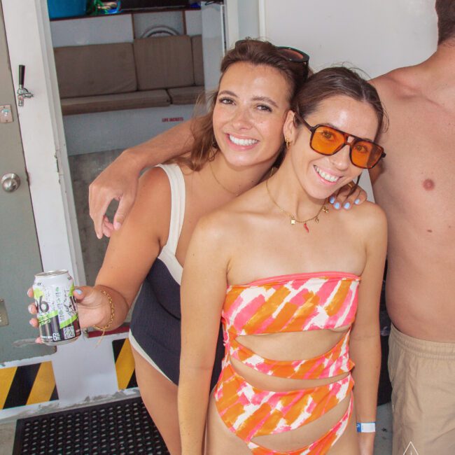 Two women in swimsuits smile at the camera on a boat. One holds a can of sparkling water, and the other wears orange sunglasses and a striped swimsuit. There is a partially visible person to the right.