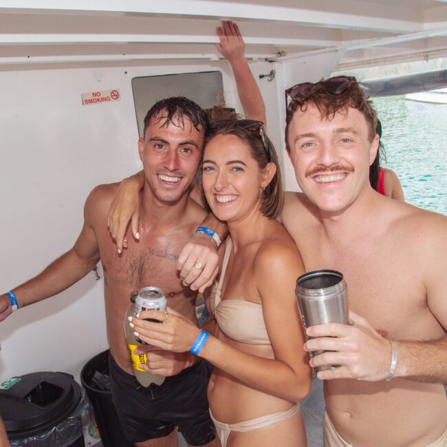 Three smiling young adults in swimsuits stand close together on a boat, holding drinks. The ocean is visible outside. They appear to be enjoying a fun, relaxed day in the sun.