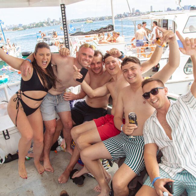 A group of six young adults in swimwear smile and cheer while posing together on a boat. The sunny scene shows other boats and people enjoying a lively day on the water in the background.