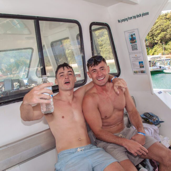 Two smiling, shirtless men sit on a boat bench, one holding a water bottle, with lush green trees and clear water visible outside the boat window. Both appear relaxed and happy, enjoying a sunny day.