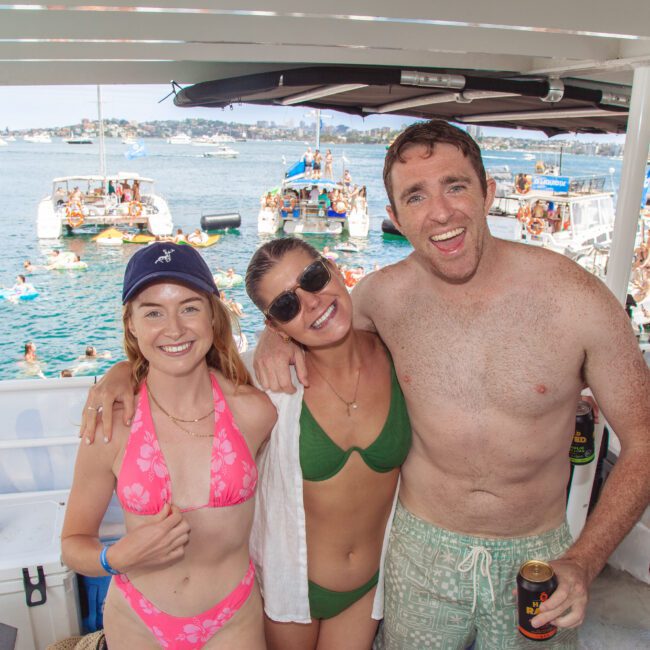 Three people in swimwear smile and pose together on a boat during a lively party; other boats and people swimming are visible in the background, with a sunny waterfront scene.