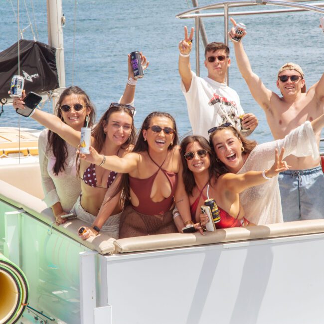 A group of young adults in swimwear smile and pose with drinks on a boat under sunny skies. Some hold up peace signs, and the ocean is visible in the background, suggesting a fun, social atmosphere.