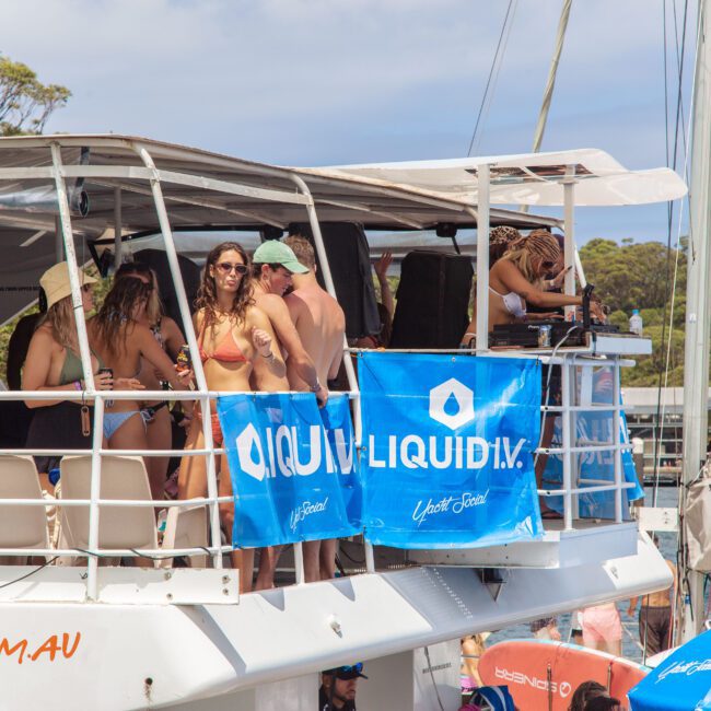 A group of people in swimsuits and sunglasses stand and socialize on the deck of a white boat, decorated with blue "LIQUID I.V." banners, docked near trees and other boats in sunny weather.