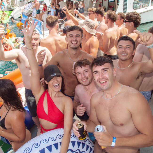 A group of smiling young adults in swimsuits pose on a boat during a lively summer party. People swim and float on inflatables in the water nearby, enjoying sunny weather and drinks.