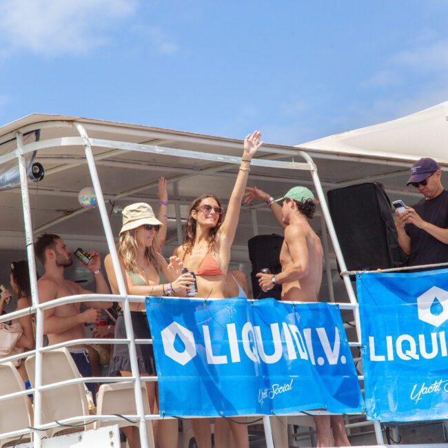 A group of young adults in swimsuits enjoy drinks and music on a boat decorated with blue "Liquid I.V." banners under a sunny sky. One woman in sunglasses waves with a smile.