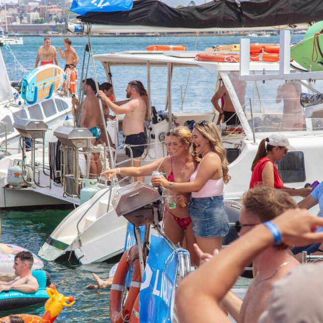 A lively scene of people enjoying a boat party, with many in swimsuits on deck and in the water on inflatables, set against a backdrop of boats and a sunny sky.