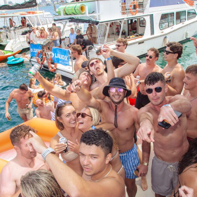 A large group of young adults in swimwear enjoy a lively boat party on the water, holding drinks, dancing, and smiling, with boats, inflatable floats, and clear blue water in the background.
