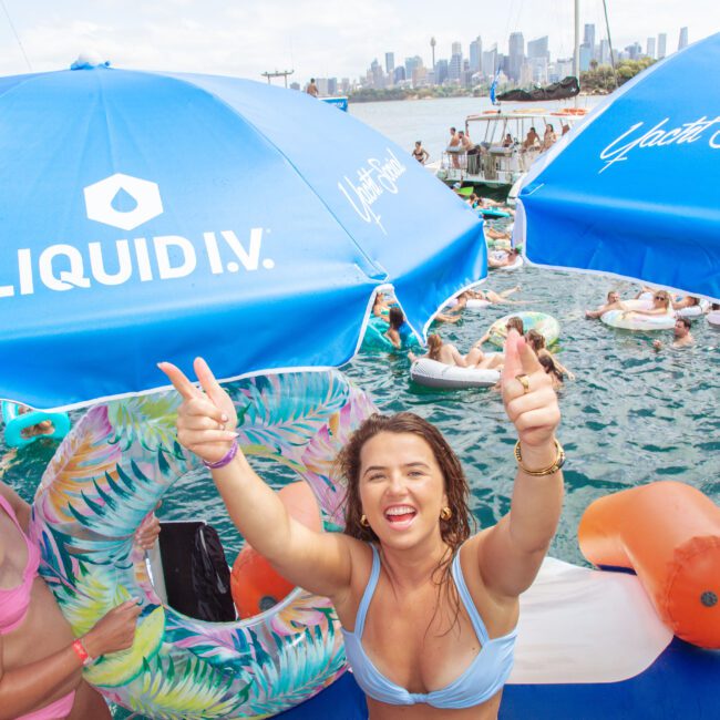 A woman in a swimsuit smiles and makes peace signs at a lively pool party, surrounded by colorful floaties and blue umbrellas with "LIQUID I.V." branding. Many people are swimming and relaxing in the water nearby.