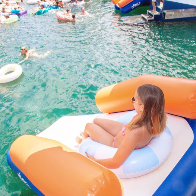 A woman in sunglasses relaxes on a large inflatable chair on the edge of a boat, overlooking a lake filled with people swimming and floating on colorful inflatables. "Yacht Social Club" logo is visible in the corner.