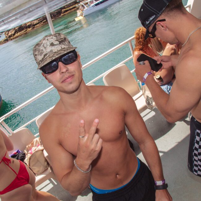 A shirtless man wearing sunglasses and a camouflage hat flashes a peace sign on a boat. Other people in swimwear are around him, and a body of water with boats is visible in the background.