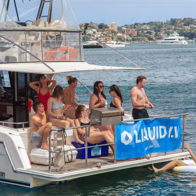 A group of people in swimwear relax and socialize on the deck of a docked yacht named "Voyager," with a "Liquid IV" banner. One man floats in the water nearby. Other boats and a shoreline are visible in the background.