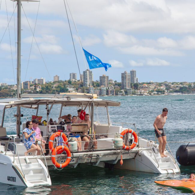 A group of people relax and socialize on a catamaran anchored on the water, with a man stepping off the boat onto a floating mat. City buildings and a partly cloudy sky are visible in the background.
